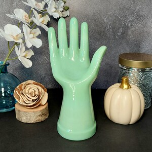 May include: A ceramic hand shaped like a ring holder, in a light green color. The hand is raised with fingers spread apart. The hand is on a black surface.