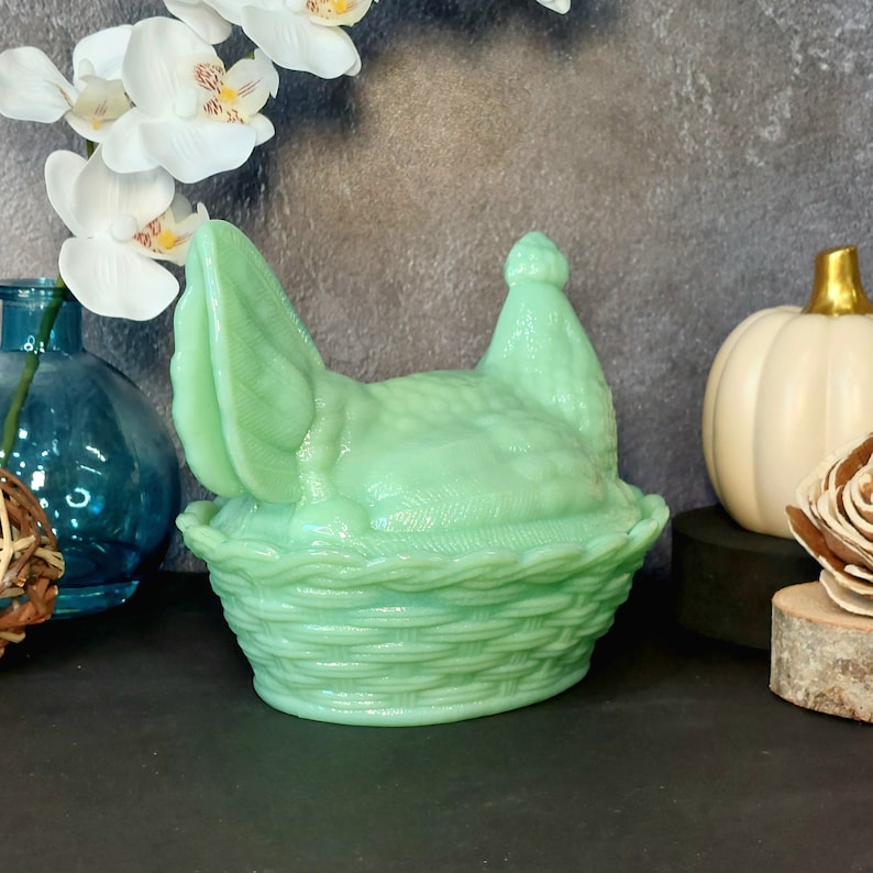 May include: A light green hen-shaped trinket box with a basket weave base. The lid features the hen's head and tail feathers. The box is made of glass and is a decorative item. The background includes a white pumpkin and a blue vase.