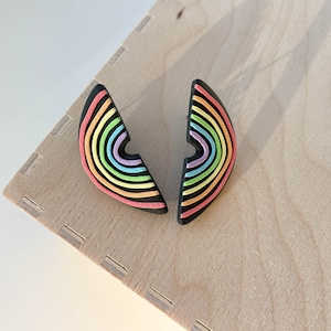 Könnte beinhalten: Ein Paar regenbogenfarbene Halbkreis-Ohrringe. Die Ohrringe sind aus schwarzem Polymerton mit einem Regenbogenmuster. Der Regenbogen besteht aus Rot, Orange, Gelb, Grün, Blau, Indigo und Violett.