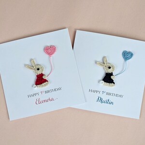 May include: Two white cards with crocheted bunny figures holding heart-shaped balloons. One bunny wears a red dress, the other a black dress. Text reads "Happy 1st Birthday" and names.