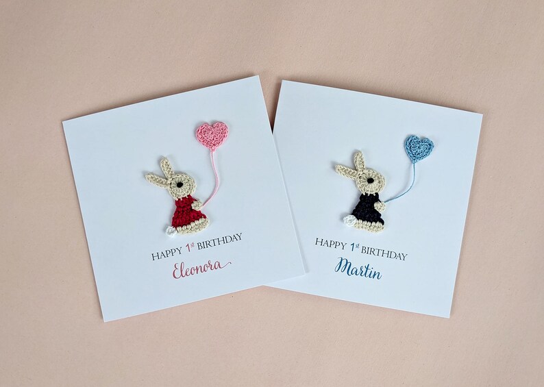 May include: Two white cards with crocheted bunny figures holding heart-shaped balloons. One bunny wears a red dress, the other a black dress. Text reads "Happy 1st Birthday" and names.