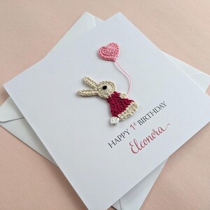 May include: A handmade birthday card with a crocheted bunny holding a pink heart balloon. The card reads "Happy 1st Birthday" and the name "Eleonora" in pink script. The card is white and comes with a white envelope.
