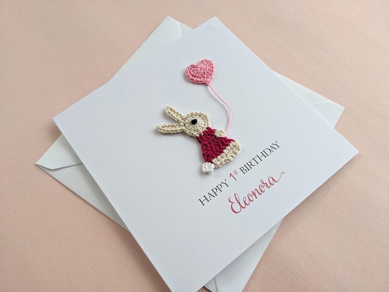 May include: A handmade birthday card with a crocheted bunny holding a pink heart balloon. The card reads "Happy 1st Birthday" and the name "Eleonora" in pink script. The card is white and comes with a white envelope.