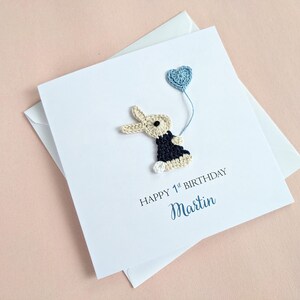 May include: A white birthday card with a crocheted bunny holding a blue heart-shaped balloon. The card reads "HAPPY 1st BIRTHDAY" and the name "Martin" in a script font. The card is on top of a white envelope.