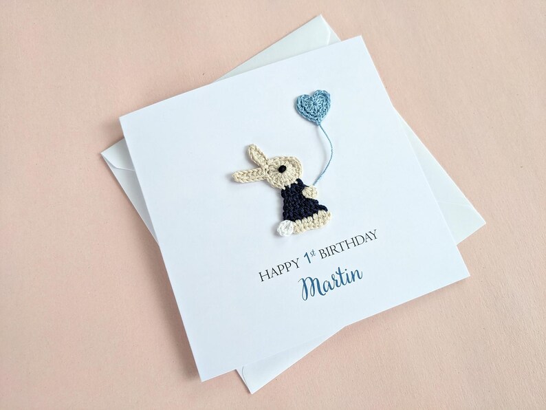 May include: A white birthday card with a crocheted bunny holding a blue heart-shaped balloon. The card reads "HAPPY 1st BIRTHDAY" and the name "Martin" in a script font. The card is on top of a white envelope.
