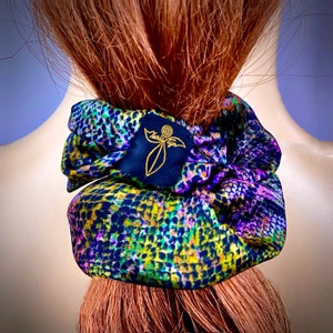 Puede incluir: Un scrunchie de pelo colorido con un logotipo negro y dorado que dice "Maria Sun". El scrunchie tiene un estampado de leopardo en tonos verdes, morados y amarillos.