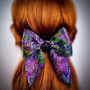 Könnte beinhalten: Eine Schleife mit violettem, grünem und schwarzem Muster und einem schwarzen Knopf in der Mitte. Die Schleife ist in den Haaren einer Person gebunden.