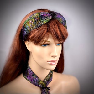 Pode incluir: Uma faixa de cabeça com padrão roxo, verde e dourado com um botão preto. A faixa de cabeça é usada em um manequim com cabelo longo e vermelho.