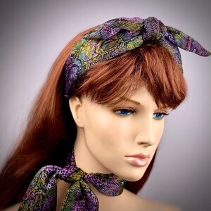 May include: A purple, green and yellow patterned headband with a knotted bow on top. The headband is worn by a mannequin with long, red hair.