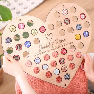 May include: A heart-shaped wooden bottle cap holder with numerous circular openings. The holder is filled with various bottle caps and features the names "David & Fiona" and the phrase "EVERY BOTTLE CAP TELLS A STORY."