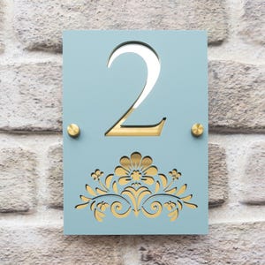 Könnte beinhalten: Hellblaues Hausnummernschild mit der ausgeschnittenen Zahl 2 und einem goldfarbenen Hintergrund. Das Schild hat unten ein goldenes Blumendesign und ist mit zwei goldfarbenen Schrauben befestigt.