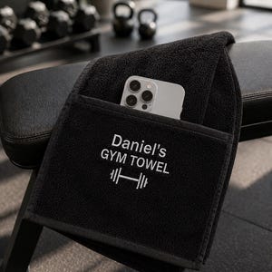 May include: A black gym towel featuring a pocket with the embroidered white text 'Daniel's GYM TOWEL' above a barbell icon. A light-colored smartphone is tucked into the pocket, resting on the textured fabric of the towel.