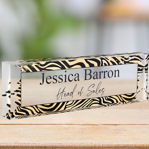Puede incluir: Una placa de nombre de escritorio de acrílico transparente con un diseño de estampado de cebra negro y dorado. La placa dice "Jessica Barron, Head of Sales."