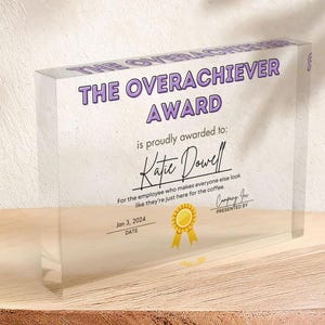 Może przedstawiać: Akrylowa tabliczka nagrody w kolorze fioletowym i białym z tekstem "The Overachiever Award" i "is proudly awarded to: Katie Dowell" oraz "For the employee who makes everyone else look like they're just here for the coffee." Tabliczka zawiera również datę "Jan 3, 2024" i tekst "Company Inc. PRESENTED BY".