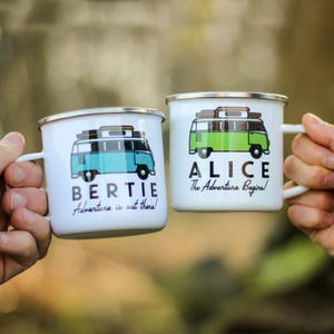 May include: Two white enamel mugs with silver rims. One mug features a blue camper van graphic with the name "BERTIE" and the text "Adventure is out there!". The other mug has a green camper van graphic with the name "ALICE" and the text "The Adventure Begins!"