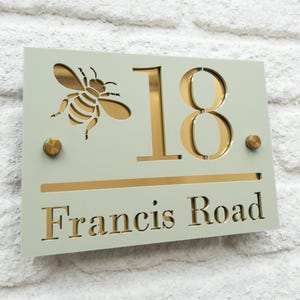 Peut inclure: Panneau de numéro de maison rectangulaire avec un fond vert clair. Il présente une découpe d'abeille dorée, le numéro "18" et les mots "Francis Road" en or. Deux rivets dorés sont également présents.