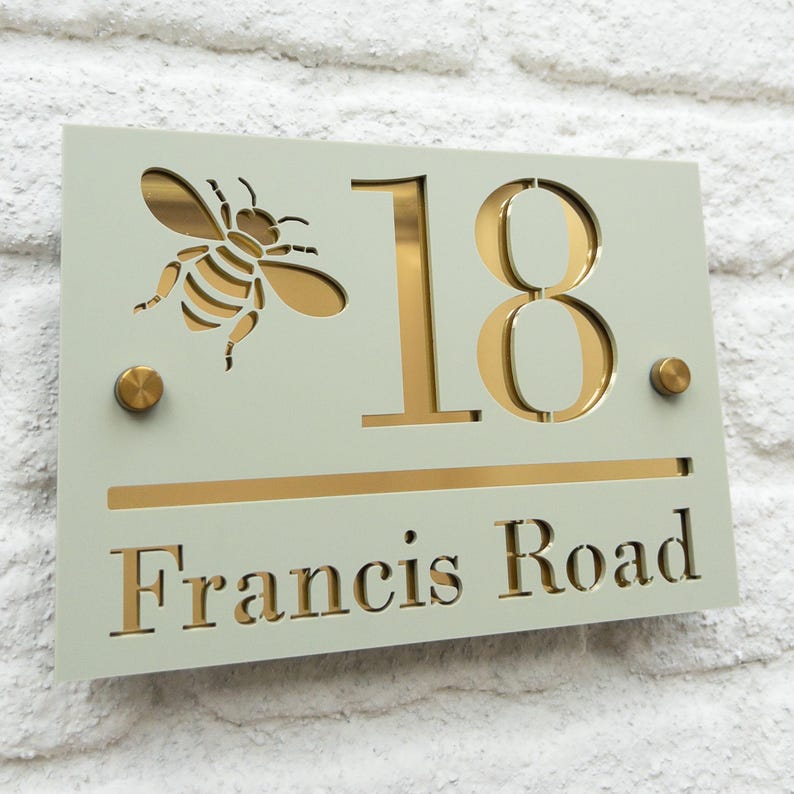 Puede incluir: Un letrero rectangular con el n&uacute;mero de la casa, con fondo verde claro. Presenta un recorte de abeja dorada, el n&uacute;mero "18" y las palabras "Francis Road" en dorado. Tambi&eacute;n tiene dos remaches dorados.