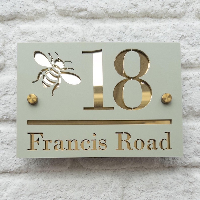 Puede incluir: Un letrero rectangular verde salvia con el n&uacute;mero 18 en dorado. Una abeja recortada est&aacute; a la izquierda, y las palabras "Francis Road" est&aacute;n en la parte inferior. El letrero tiene detalles dorados y tachuelas color lat&oacute;n.