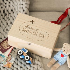 Puede incluir: Una caja de madera clara con el texto "Archie's ADVENTURE BOX" y un pequeño avión. Un coche de juguete de madera, un oso de peluche y un pasaporte están al lado de la caja. La caja tiene una cinta roja.