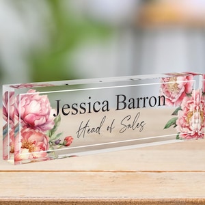 May include: A clear acrylic desk nameplate with a floral design featuring pink peonies. The nameplate reads "Jessica Barron, Head of Sales".