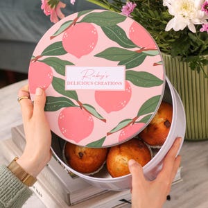 May include: A round, white tin with a peach and leaf design on the lid, featuring the text "Ruby's Delicious Creations." The tin is open, revealing several muffins. The lid is being held open by a person.