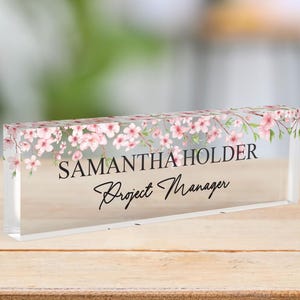 Könnte beinhalten: Ein klares Acryl-Namensschild für den Schreibtisch mit einem Blumendesign aus rosa Blüten und grünen Blättern. Das Namensschild lautet "SAMANTHA HOLDER Project Manager".