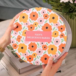 May include: A round tin featuring a floral design with orange, yellow, and pink flowers on a white background. The tin has a pink label that reads "NAN'S DELICIOUS BAKES". The tin is held by a person.