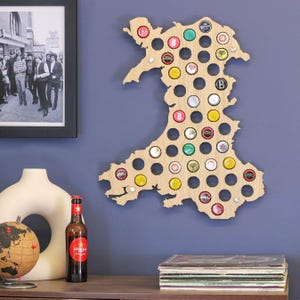 May include: Wooden bottle cap map of Wales, filled with various beer bottle caps. The map is mounted on a blue wall. A framed black and white photo and a bottle of beer are visible in the background.