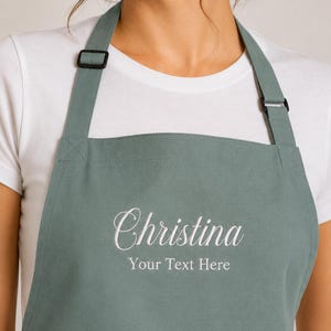 May include: Sage green apron embroidered with the name "Christina" in white script, and the text "Your Text Here" below. It features adjustable straps with black buckles. The apron is made of a soft, durable fabric, suitable for kitchen use.