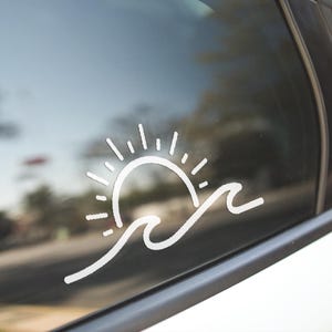May include: White vinyl decal of a sun setting over ocean waves, applied to a car window. The design features a stylized sun with radiating lines and two curved wave lines. The decal is a simple, clean design.