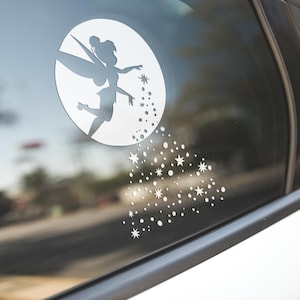 May include: A white vinyl decal of Tinkerbell, a fairy with wings, flying out of a circle with a trail of stars behind her. The decal is on a car window.