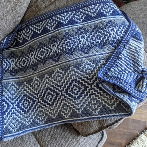 Op de afbeelding: Een blauwe en grijze gehaakte deken met een geometrisch patroon. De deken is gemaakt met verschillende steken en heeft een gefranst randje.