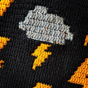 Op de afbeelding: Gehaakt stof met een grijze wolk en gele bliksemschichten op een zwarte achtergrond.