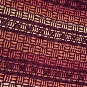 Op de afbeelding: Een close-up van een gebreide stof met een geometrisch patroon in tinten van bordeauxrood, oranje en crème.