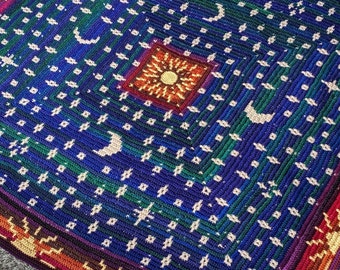 Puede incluir: Una manta de ganchillo con fondo azul y un borde colorido. La manta presenta un patrón repetitivo de estrellas, lunas y soles.