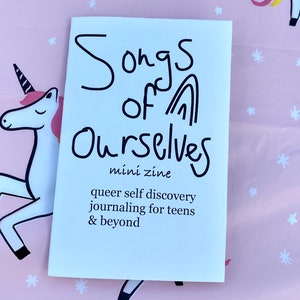 Könnte beinhalten: Ein weißes Mini-Zine mit dem Titel "Songs of Ourselves" und dem Untertitel "queer self discovery journaling for teens & beyond".