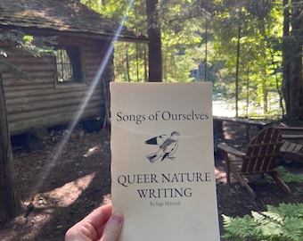 May include: A book titled "Songs of Ourselves: Queer Nature Writing" by Sage Mitchell, with a bird illustration on the cover, is held in front of a wooden cabin in a forest setting.