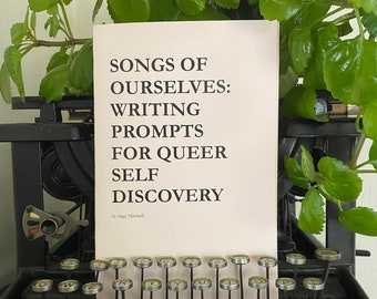 May include: A book titled "Songs of Ourselves: Writing Prompts for Queer Self Discovery" by Sage Mitchell, resting on a vintage typewriter.
