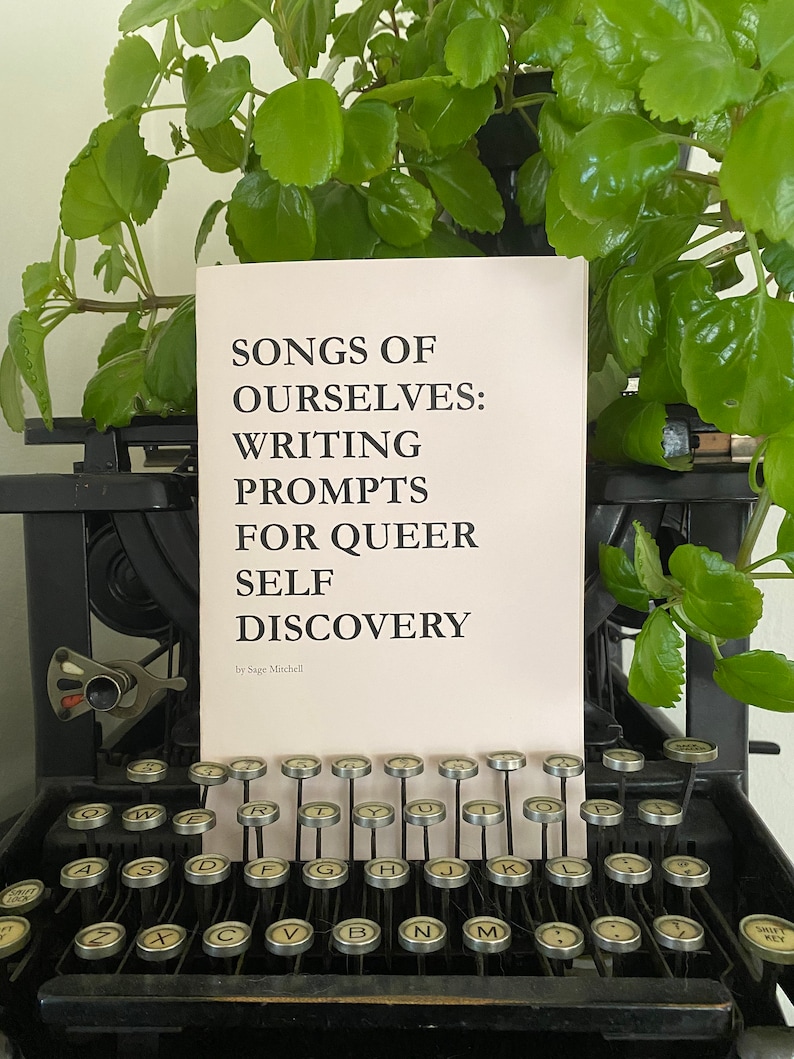 May include: A book titled "Songs of Ourselves: Writing Prompts for Queer Self Discovery" by Sage Mitchell, resting on a vintage typewriter.