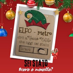 May include: A wooden Christmas elf meter with a green and red elf hat design. The text on the meter reads "ELFO - metro" and "SEI STATO". The meter has a sliding scale to indicate if someone has been "bravo" or "monello".