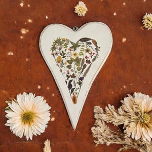May include: White wooden heart-shaped ornament with a pressed flower design. The flowers are arranged in a bouquet shape and include various colors and textures.
