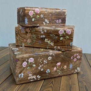 May include: Three wooden boxes, each with a floral design of pink roses, bluebells, and wildflowers. The boxes are stacked, and the wood has a warm brown colour. The boxes are made of wood.