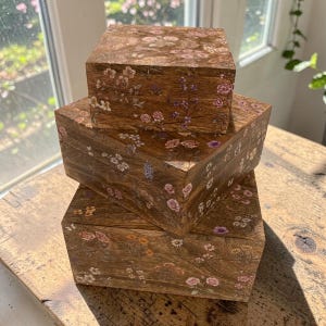 Peut inclure: Une pile de trois boîtes carrées en bois de tailles différentes, chacune présentant une finition en grain de bois brun naturel ornée d'un motif floral délicat de petites fleurs roses, blanches, jaunes et violettes avec des tiges vertes.