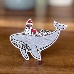 May include: A gray whale with a white lighthouse and red roof on its back. The whale is embroidered with white thread and has a small paper boat on its back.
