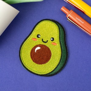 May include: A cheerful avocado patch with a smiling face, embroidered in shades of green, brown, and pink, sits on a blue surface. Two pens, one yellow and one orange, are positioned nearby. A green plant adds a touch of freshness to the scene.