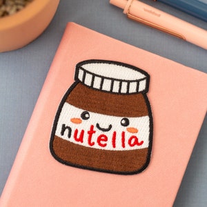 May include: A pink notebook with a brown and white embroidered Nutella jar patch. The jar has a smiling face and the word "Nutella" is embroidered in red.