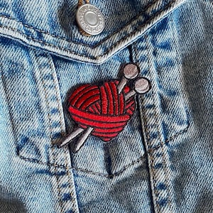 Könnte beinhalten: Ein roter herzförmiger Wollknäuel mit zwei Stricknadeln, die herausragen. Der Wollknäuel und die Nadeln sind auf eine blaue Jeansjacke gestickt.