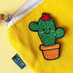 Peut inclure: Pochette zippée en toile jaune avec un cactus brodé vert surmonté d'une fleur rouge. Le cactus sourit et a un pot brun. La pochette porte une étiquette verte avec le texte "GIS".