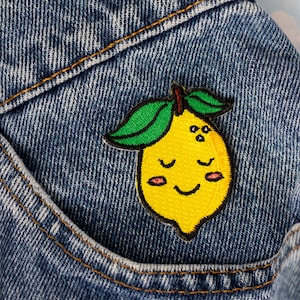 May include: A yellow lemon patch with a smiling face, green leaves, and a brown stem is sewn onto a blue denim pocket. The patch is a fun, colorful accessory.