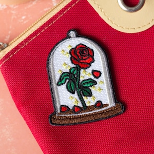 Peut inclure: Une rose rouge avec des feuilles vertes et une tige dorée est enfermée dans un dôme de verre avec une base brune. Le dôme est décoré d'étoiles dorées et de cœurs rouges. Le patch est sur un fond de tissu rouge.